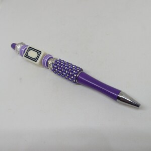 May include: A purple ballpoint pen featuring a silver-toned metal tip and a barrel section adorned with small purple rhinestones. The upper portion includes a square white bead with a black border design, flanked by purple and white striped beads.