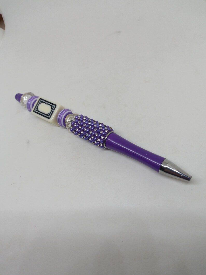 May include: A purple ballpoint pen featuring a silver-toned metal tip and a barrel section adorned with small purple rhinestones. The upper portion includes a square white bead with a black border design, flanked by purple and white striped beads.