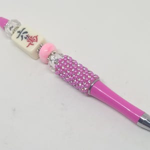 May include: A pink ballpoint pen featuring a decorative top section with a white mahjong tile displaying red and black characters, accented by clear faceted beads, a solid pink bead, and a cylindrical barrel covered in small pink and clear rhinestones.