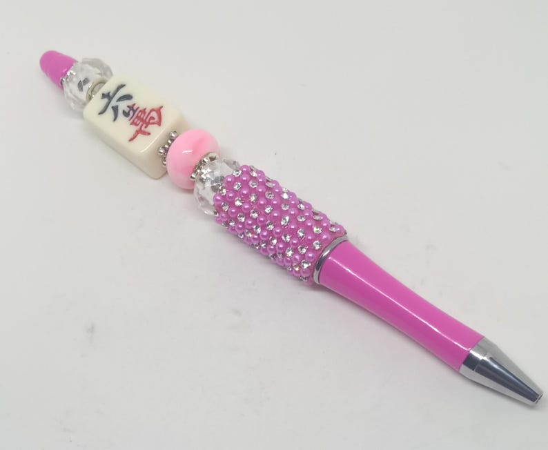 May include: A pink ballpoint pen featuring a decorative top section with a white mahjong tile displaying red and black characters, accented by clear faceted beads, a solid pink bead, and a cylindrical barrel covered in small pink and clear rhinestones.