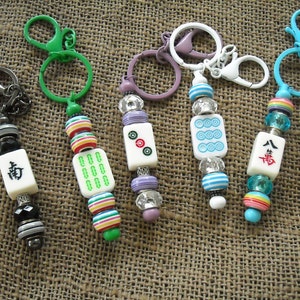 May include: Five colorful keychains with different charms. Each keychain features a white tile with Chinese characters and a variety of colorful beads. The charms include a green tile with a circle pattern, a blue tile with a circle pattern, a tile with a red circle and two dots, a tile with a black character, and a tile with a red character.
