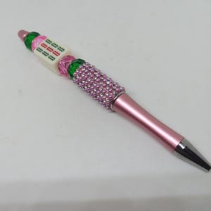 May include: A pink ballpoint pen featuring a metallic pink barrel and a silver tip. The upper section is decorated with a cluster of small pink pearls, followed by a series of green and pink faceted beads and a white cube bead with green text.
