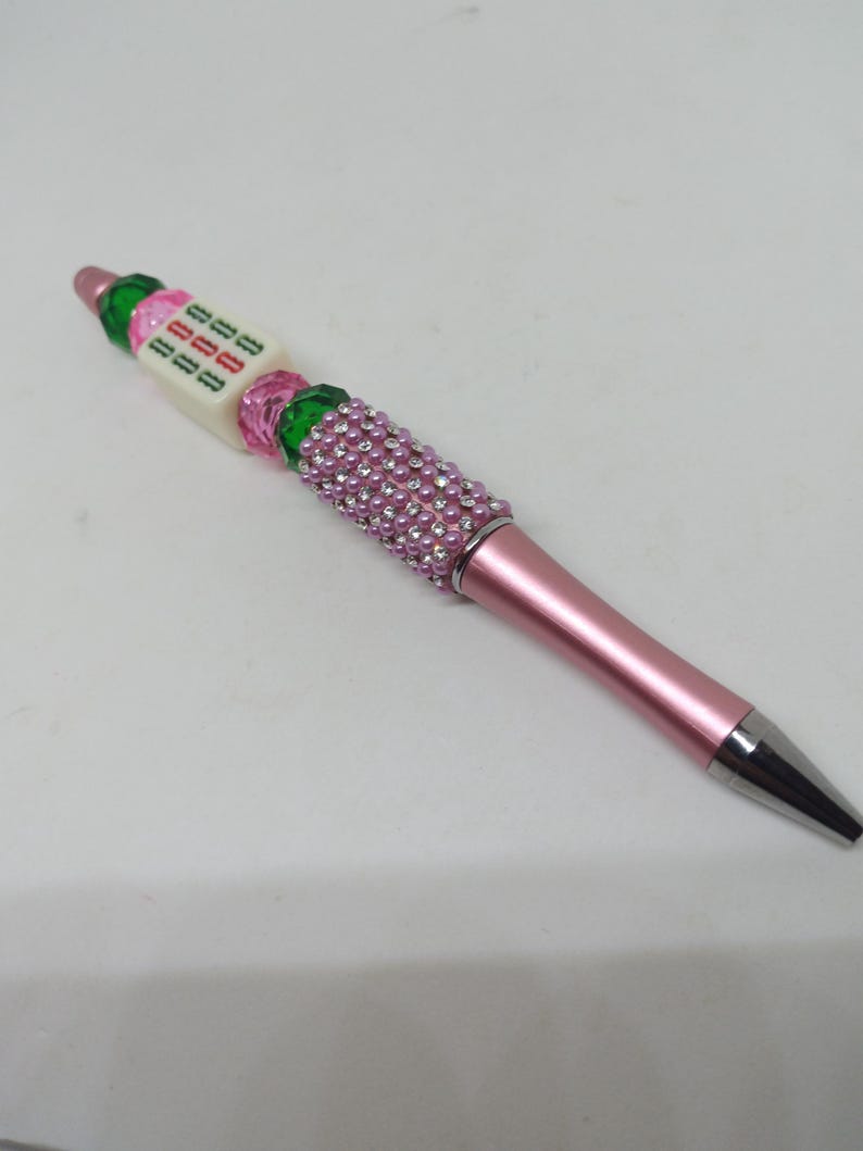 May include: A pink ballpoint pen featuring a metallic pink barrel and a silver tip. The upper section is decorated with a cluster of small pink pearls, followed by a series of green and pink faceted beads and a white cube bead with green text.