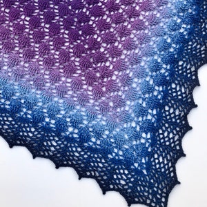 Puede incluir: Un chal de ganchillo con un degradado de colores morado, azul y blanco. El chal tiene un delicado patrón de encaje y un borde festoneado.