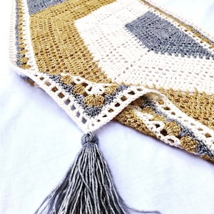 May include: A crocheted throw blanket with a geometric pattern in shades of white, gold, and grey. The blanket has a fringe tassel on one corner.