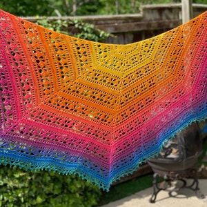 May include: A rainbow ombre crochet shawl with a lacy pattern. The shawl is hanging in a garden setting.