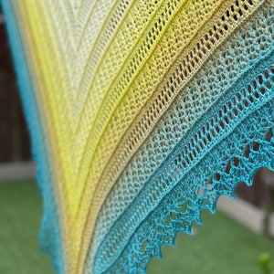 Puede incluir: Un chal de ganchillo con un degradado de colores amarillo, verde y azul. El chal está hecho con un delicado patrón de encaje.