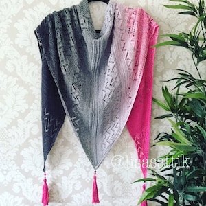 May include: A gray and pink triangular shawl with a lacy pattern and tassels. The shawl is made of yarn and has a gradient color pattern. The shawl is hanging on a wall with a floral pattern.