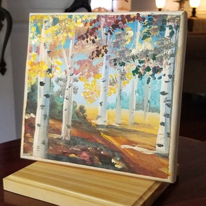 May include: A painting on canvas of a forest scene with birch trees in the fall. The trees have yellow, orange, and red leaves. The background is a blue sky with white clouds. The painting is on a wooden easel.