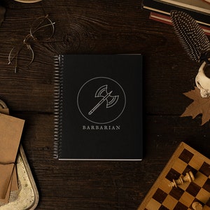 Puede incluir: Un cuaderno negro con espiral con un gráfico de hacha blanco y la palabra "BARBARIAN" en la portada. El cuaderno está sobre una superficie de madera oscura, rodeado de un tablero de ajedrez, libros y una calavera.