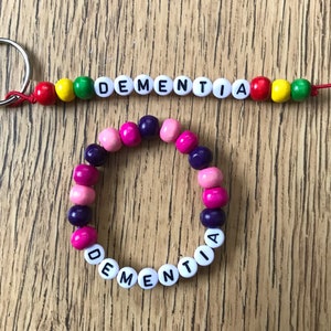 Peut inclure: Un bracelet en perles blanches avec le mot "DEMENTIA" &eacute;crit en lettres noires. Le bracelet est compos&eacute; de perles roses, violettes et blanches. Un porte-cl&eacute;s assorti avec le m&ecirc;me mot est &eacute;galement pr&eacute;sent&eacute;.