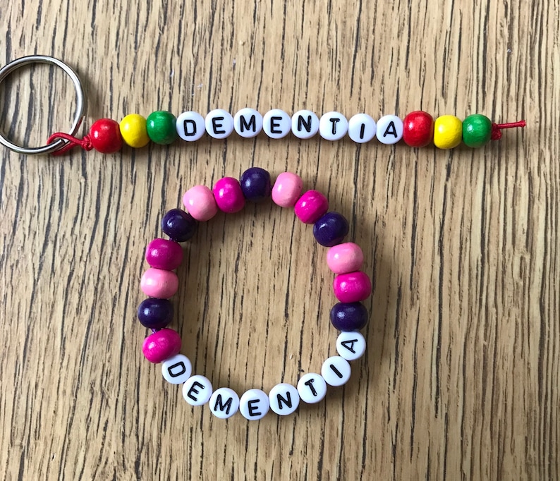 Peut inclure: Un bracelet en perles blanches avec le mot "DEMENTIA" &eacute;crit en lettres noires. Le bracelet est compos&eacute; de perles roses, violettes et blanches. Un porte-cl&eacute;s assorti avec le m&ecirc;me mot est &eacute;galement pr&eacute;sent&eacute;.