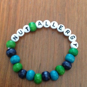 Peut inclure: Un bracelet en perles de bois avec les mots "NUT ALLERGY" &eacute;crits en lettres blanches sur des perles noires. Les perles sont vertes, bleues et turquoise.