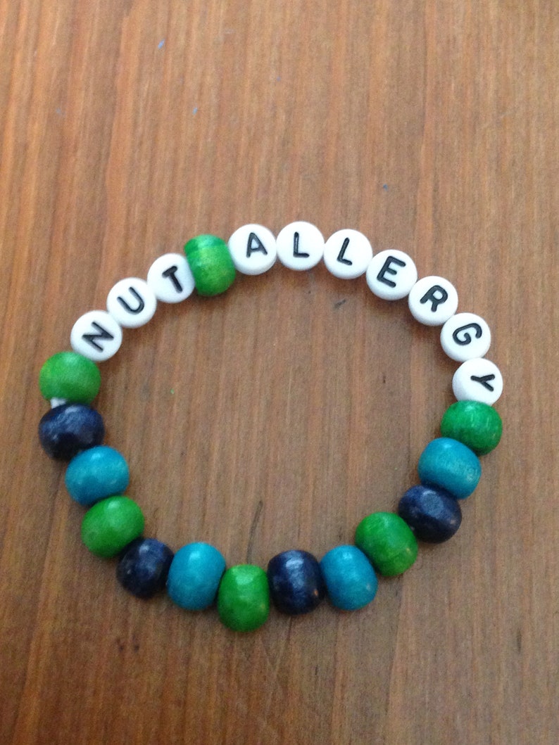Peut inclure: Un bracelet en perles de bois avec les mots "NUT ALLERGY" &eacute;crits en lettres blanches sur des perles noires. Les perles sont vertes, bleues et turquoise.