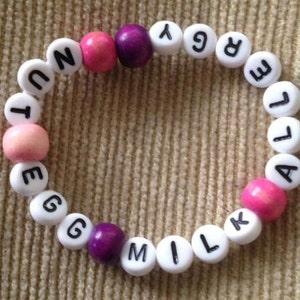 Peut inclure: Un bracelet en perles blanches avec les mots "EGG MILK ALLERGY" &eacute;crits en lettres noires. Les perles sont entrecoup&eacute;es de perles roses et violettes.