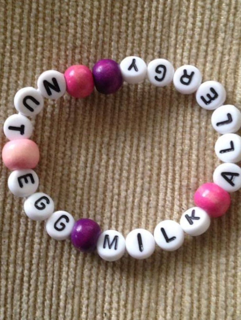 Peut inclure: Un bracelet en perles blanches avec les mots "EGG MILK ALLERGY" &eacute;crits en lettres noires. Les perles sont entrecoup&eacute;es de perles roses et violettes.
