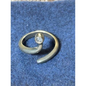 May include: A silver-toned bypass ring featuring a smooth, open-ended band that curves into a spiral shape. At one end of the band, a single, small, clear, round-cut stone is set in a circular metal bezel, creating a simple and clean design.