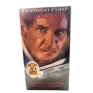 May include: A VHS tape of the movie "Air Force One" starring Harrison Ford. The cover features a close-up of Harrison Ford's face and the title in bold red letters. A Columbia Pictures logo is visible, along with a quote about the film.