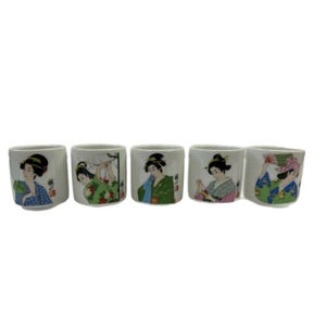 May include: Five white ceramic sake cups. Each cup features a different illustration of a geisha in traditional attire, with vibrant colors and intricate details. The cups are arranged in a row, showcasing the variety of designs.