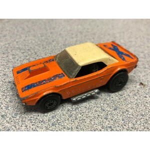 May include: Orange toy car with a cream-colored roof and blue stripes. The toy car has black wheels and a silver exhaust pipe. The word "Dodge" is visible on the side of the car.