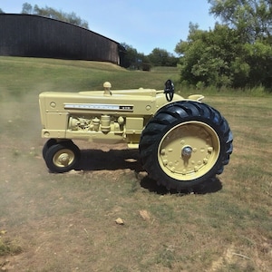 May include: A vintage Cockshutt 570 tractor in a field. The tractor is painted a light yellow color with black tires. The tractor has a black steering wheel and is parked on a grassy area.
