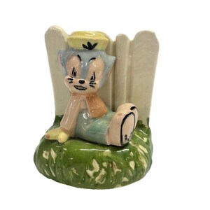 May include: A vintage ceramic figurine of a cartoon cat wearing a yellow hat, orange scarf, and green overalls, seated against a white fence. The figurine is on a green grassy base, with a charming, nostalgic aesthetic.