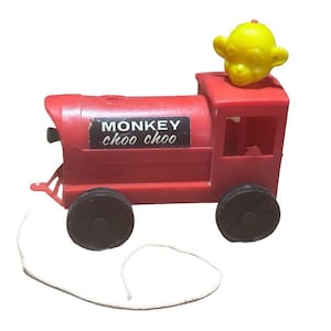 May include: A red toy train with black wheels and a yellow monkey head on top. The train has the words "MONKEY choo choo" on the side and a white string attached. A classic pull-along toy.