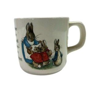 May include: A white ceramic mug featuring a colorful illustration of four rabbits. One larger rabbit wears a blue coat, while three smaller rabbits wear red coats. The mug includes printed text on the side and has a curved handle on the right side.