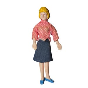 May include: A vintage doll with a yellow bob hairstyle, wearing a pink polka-dot blouse, a denim skirt, and blue flats. The doll has a pale complexion and is standing upright.