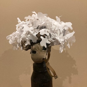 May include: A military helmet covered in white camouflage netting and leaf-shaped cutouts. The helmet is mounted on a dark, cylindrical object, possibly a gas cylinder. A tan strap hangs from the helmet.