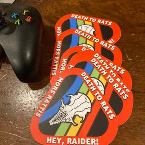 May include: A stack of red, black, and rainbow-colored stickers. Each sticker has the text "DEATH TO RATS" and "HEY, RAIDER!" with a skull graphic. The stickers are on a wooden surface.