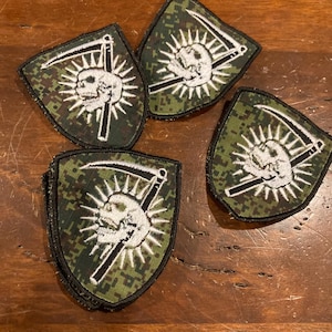 May include: Four shield-shaped patches with a skull and scythe design. The patches feature a white skull with radiating lines, a black scythe, and a green and brown camouflage background. The patches are likely for clothing or accessories.