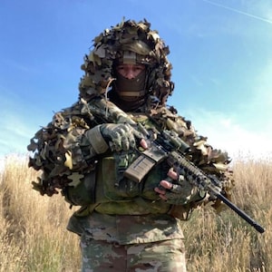 May include: A tactical uniform featuring a ghillie hood with artificial oak leaves in shades of brown and green. The gear includes a dark face mask, camouflage pants, and a rifle with a tan magazine and black barrel held in gloved hands.