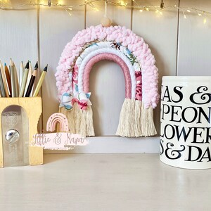 May include: A pink and white rainbow wall hanging with a floral pattern and tassels. The rainbow is made of yarn and fabric. The text "Little & Nana" is written on a white surface below the rainbow.