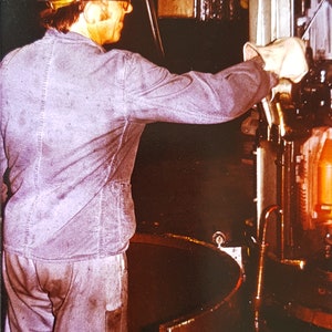 May include: A worker wearing a safety helmet and face shield operates a large industrial machine. The machine has a metal casing and a glowing orange interior.