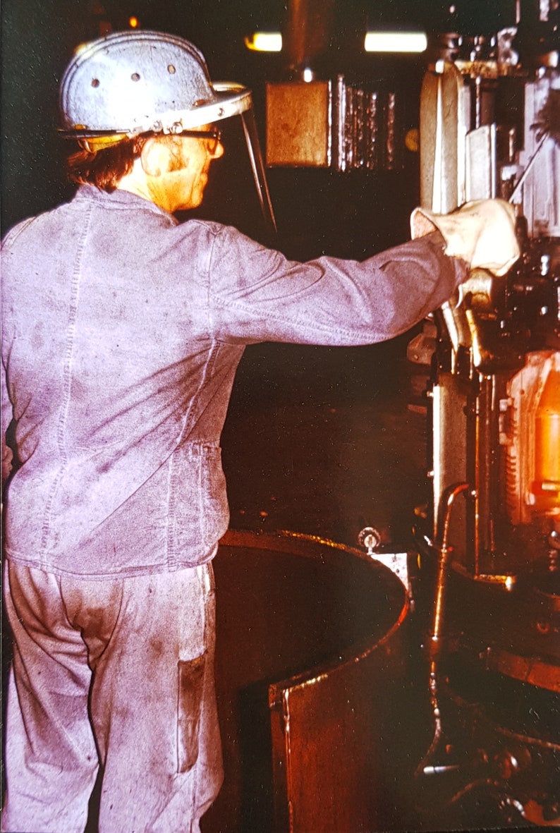May include: A worker wearing a safety helmet and face shield operates a large industrial machine. The machine has a metal casing and a glowing orange interior.