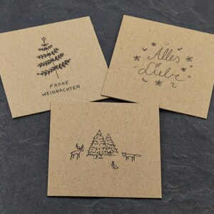 May include: Three brown kraft paper cards with black ink illustrations. The first card says "Frohe Weihnachten" and has a stylised Christmas tree. The second card says "Alles Liebe" with a heart and flowers. The third card has a stylised forest scene with a deer, a fox, and a rabbit.