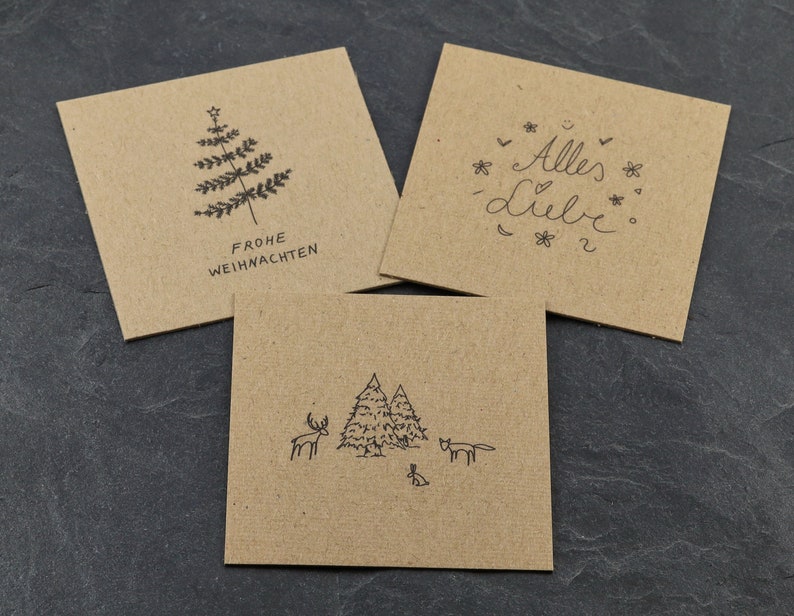May include: Three brown kraft paper cards with black ink illustrations. The first card says "Frohe Weihnachten" and has a stylised Christmas tree. The second card says "Alles Liebe" with a heart and flowers. The third card has a stylised forest scene with a deer, a fox, and a rabbit.