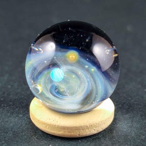 Può includere: Una biglia di vetro sferica caratterizzata da un design a nebulosa vorticosa blu scuro e nero con piccole sfere incastonate di colore giallo e azzurro simili a pianeti. La biglia misura 21 mm e poggia su un piccolo supporto in legno chiaro.