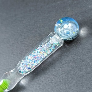 May include: A clear glass wand with a blue and white orb at the top, resembling a galaxy. The wand's handle is filled with iridescent glitter and has a green, textured base. The wand is a unique decorative item.