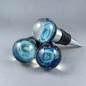May include: Three glass bottle stoppers with a blue and white galaxy design. The stoppers are round and have a silver metal top.