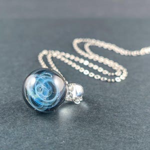 Op de afbeelding: Een bolvormige glazen hanger aan een ketting met een donkerblauwe en zwarte basis met wervelende witte en lichtblauwe nevelachtige patronen aan de binnenkant. De hanger is bevestigd aan een zilverkleurige metalen ketting met kleine ovale schakels.