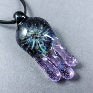 May include: A handmade glass pendant with a galaxy design. The pendant is a mix of dark blue, purple, and white colors, with a black cord. The glass has a unique shape with three rounded tips, and a black loop for the cord.