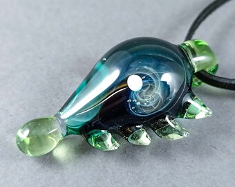 May include: A handcrafted glass pendant with a unique design. The pendant features a blend of teal, dark blue, and white colours, with a swirling pattern inside. It has a green tip and accents, and is strung on a black cord.