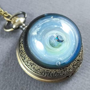 May include: A vintage-style pocket watch with a celestial design. The watch face features a swirling blue and white galaxy with a crescent moon. The watch has a gold-colored case with ornate detailing and a matching chain.