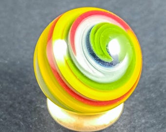 May include: A colorful glass marble with a swirl design. The marble features bands of yellow, red, white, and green, creating a spiral pattern. It sits on a small, gold-colored base, showcasing its vibrant colors and intricate design.
