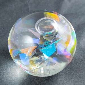 Könnte beinhalten: Ein klarer Glasbriefbeschwerer mit farbenfrohen, irisierenden Scherben im Inneren. Die Scherben reflektieren Farbtöne von Blau, Orange und Gelb. Der Briefbeschwerer ist rund und liegt auf einer dunklen Oberfläche.