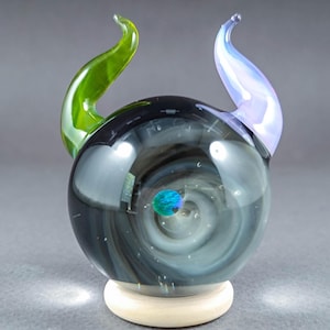 May include: A handcrafted glass sculpture featuring a swirling, galaxy-like interior with a small, colorful orb at the center. The sculpture has two curved horns, one green and one lavender, and sits on a light-colored wooden base.