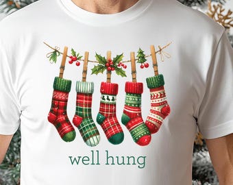 May include: White t-shirt with a Christmas design of stockings hanging on a string, with the text "well hung" in green. The stockings are red and green with various patterns. Christmas-themed apparel.