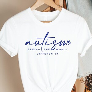 May include: White t-shirt with the word "autism" in blue script, with a heart-shaped dot over the "i". Below, the text reads "Seeing the world differently". The shirt is hanging on a wooden hanger.
